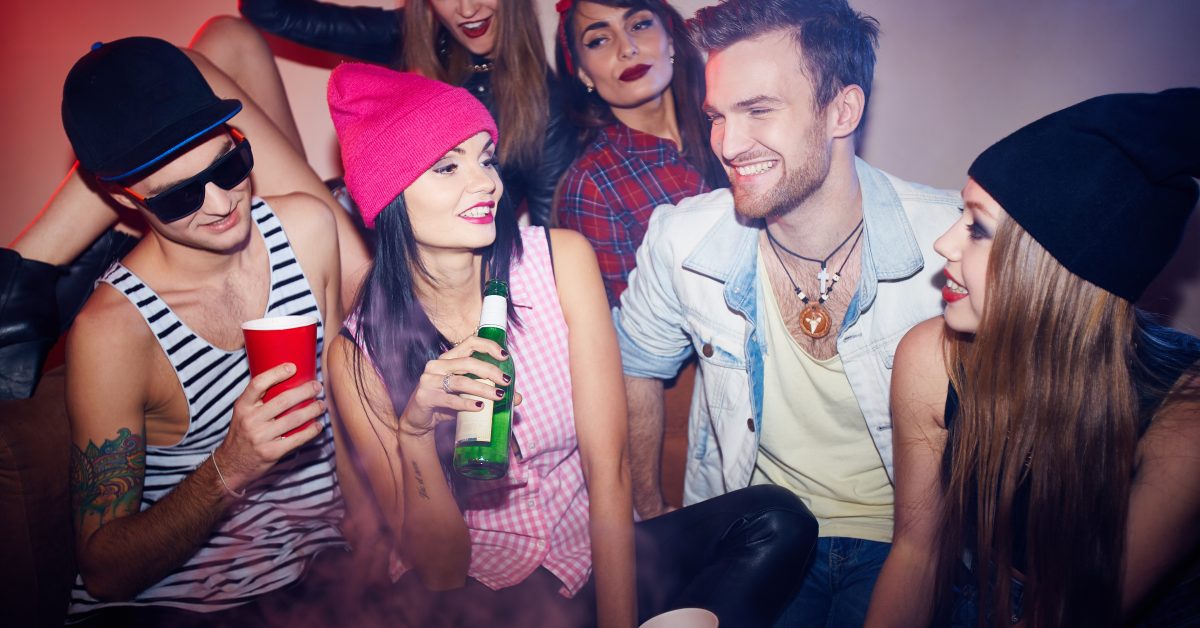 Stylish men and women enjoying raging swag party, talking and laughing on sofas with beer in crowded smoky room